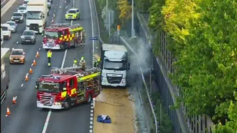 A lorry parked in a motorway layby ihas smoke emitting from it. Two fire engines are parked nearby.