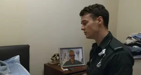 Stephen Doohan sits by a bedside wearing green surgical gloves. He has short brown hair and looks down. He is wearing a green NHS paramedic uniform
