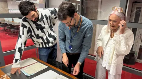 Two men and a woman look at documents on a table.