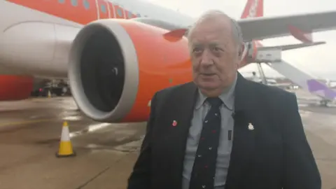 Ant Saddington/BBC An older man is smartly dressed and standing in front of an orange and white plane at an airport.