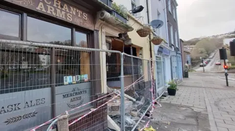 The outside of a pub after a car collided with it. There is a metal fence blocking the entrance, and there is debris in front of the building.