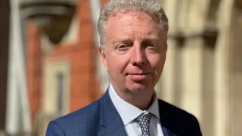 Alex Allinson is wearing a navy suit, light blue shirt and spotted tie. He's standing in front of a brick building and has short fair hair.