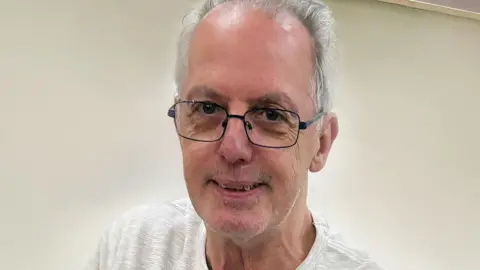 Man in glasses with grey hair balding at front looks at camera with slight smile, head and shoulders shot wearing white t shirt.