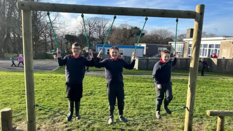 WILLASTON SCHOOL Three primary school boys stand on a rope balancing beam in a playing field while holding on to rope handles, with the school behind them.