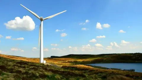 A single white wind turbine in an isolated area of the countryside, with blue skies and some clouds and a lake close to the structure.