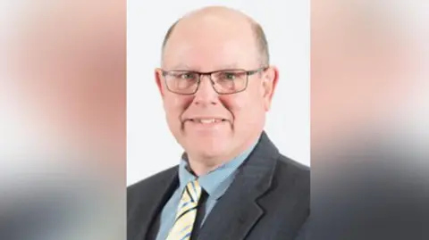 Westmorland and Furness Council A portrait image of a smiling Andrew Jarvis. he has grey hair around his temples and glasses. his eyes are blue and he wears a blue shirt, yellow tie and a grey suit jacket. 