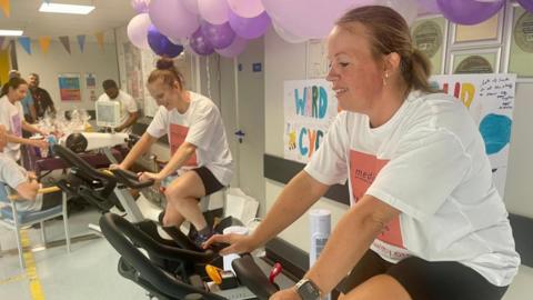 Two members of hospital staff dressed in sponsored T-shirt pedalling on exercise bikes in a corridor at Leighton Hospital