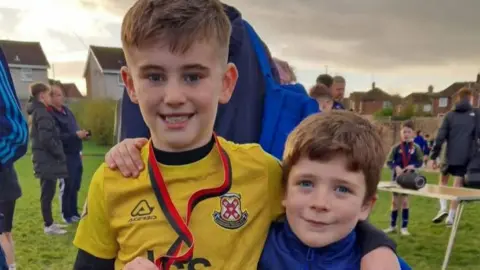 Kim Holmes (L-R) Calvin Holmes is wearing a yellow football kit and holding a medal and Reuben McLaughlin