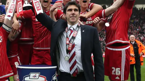 Middlesbrough boss Aitor Karanka, wearing a dark suit jacket, white shirt and red and white striped tie, lifts the promotion trophy aloft with his right hand after Boro won promotion to the Premier League in May 2016.