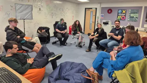 A group of people in a circle having a discussion. Young people sat and look towards someone speaking while councillor Emma Bushell sits with her arms on her legs and listens intently.