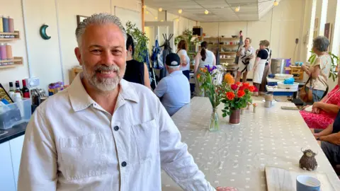 A man with grey hair and a grey beard in a white collared shirt stood in front of a pottery class.