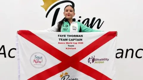 Jodie Thorman Faye holding a flag that reads "Faye, team captain, Dance World Cup 2026, Team N.Ireland" The flag has the logos of two sponsors, and her dance academy's badge on it, titled "Reign". She is standing Infront of a white wall that has her dance academy's badge across it in a large print. She has brunette hair, wearing black leggings with a Dance World Cup zip up jacket. Her brunette hair is in a ponytail and she is wearing red lipstick.