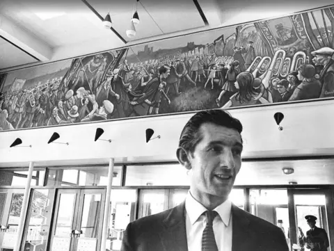 The Story/Durham County Council Norman Cornish, a thin and clean shaven man with black hair, stands in front of a large mural depicting crowds of people a the Durham Miners' Gala