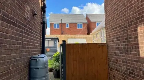 A gap in between two red brick houses, with a row of houses in the background. There is a fence panel and two water butts in the foreground. 