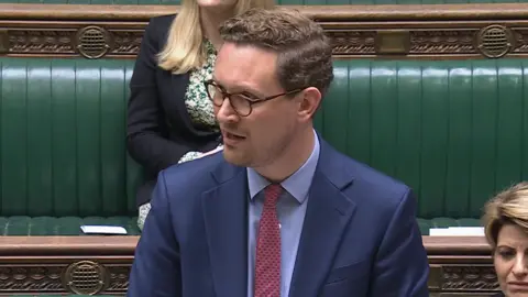 Darren Jones wearing a red tie, light blue shirt and dark blue suit, standing at the dispatch box of the House of Commons' green benches. He has twp female colleagues sitting to the right of him and behind.