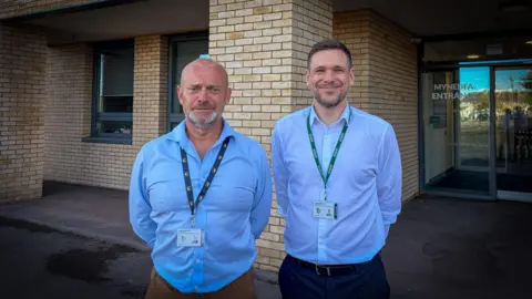two men in light blue button down shirts stand beside each other, hands behind their backs looking at the camera. they both have lanyards and in front of a beige brick school building near the glass doored entrance