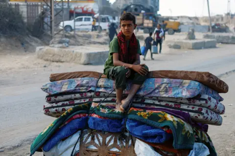 Reuters A young boy sits on a cart strapped with mattresses, blankets and other luggage. He is on a dusty road. 