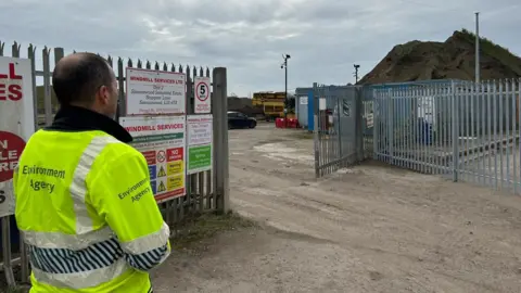 A man in a high-visibility yellow jacket with Environment Agency written on the back is standing at a site entrance next to metal fencing and looking at a large mound of waste.