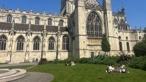 A photograph of a gothic cathedral taken on a sunny day. Two groups of people sunbathe in front.