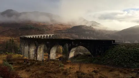 Dougie Thomas A misty day with a large, dark, rail bridge going through the centre of the image. The grass and plants in the foreground are weathered and green and yellow. Mist is covering the mountain in the background. The sky is white and grey with small points of blue poking through.