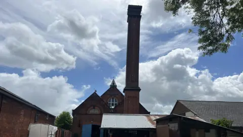General view of the Coleham Pumping station. It's a Victorian building with a tall chimney and blue doors. A few cars are parked in the yard at the front and a smaller shabby building is in the foreground