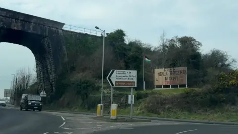 A picture of a bridge beside it is a road, road sign, Palestine flag and billboard that says FREE NIALL SHEERIN NOW! IRPWA. Driving on the road is a black vana nd silver van