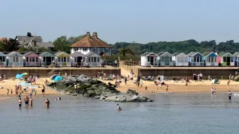 BBC Weather Watchers/Southern Broads The coast at Southwold from the sea. People are wading in the blue water and walking on the yellow sands. Above the beach is a row of colourful beach huts, trees, two roofs and pale blue sky. 