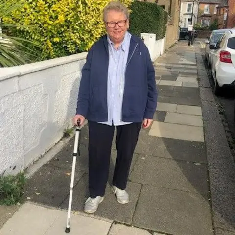 Hazel Kelly Hazel Kelly is walking on  a thoroughfare  with the assistance  of a achromatic  walking stick. She is wearing glasses and a bluish  overgarment   and airy   bluish  garment  and achromatic  trousers.