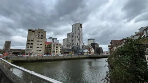 Shaun Whitmore/BBC A town waterfront area on a cloudy day. Tall buildings are lined along a body of water.
