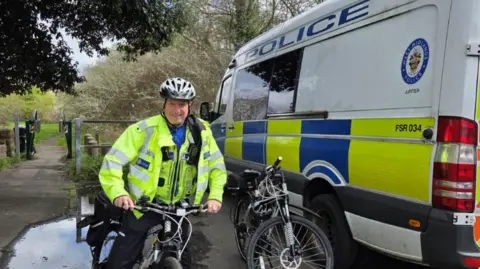West Midlands Police A police officer in a hi-vis jacket is on a bike and wearing a helmet. Trees and grass are in the background on the left and a police van is on the right.
