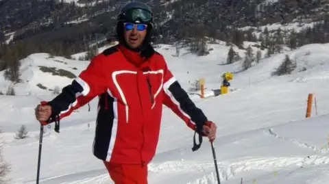 Kristian Shearan Kristian is pictured on a ski slope, wearing a red ski suit and holding ski poles. He is wearing a helmet and sunglasses and behind him is a snow-covered ski slope and trees.
