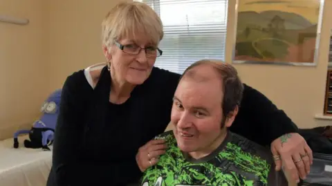 A woman with short blonde hair and black glasses, wearing a black jumper is seen with her arms around a man who is sitting down. The man has brown hair, thinning on top, and is wearing a green and black sports top.