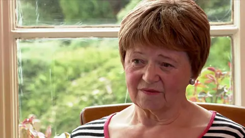 Joan Lawrence, Claudia's mother, looks to the left of camera. She is wearing a stripey top and is sitting in a chair in front of a window with plants outside the window behind her.