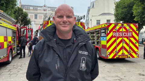 Deputy Chief Fire Officer Bryn Coleman smiles at the camera, there are fire engines behind him
