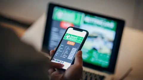 A phone is being held in front of a blurry person and it shows a gambling website. There is a laptop showing a blurry gambling screen in the background. 