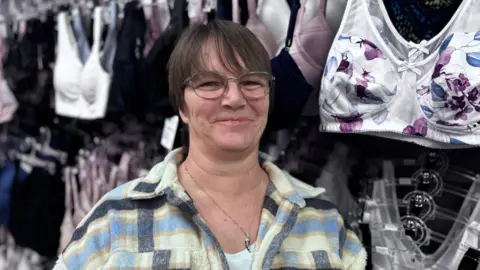 BBC Woman with short brown hair and glasses, wearing a checked shirt, smiling at the camera in a lingerie shop. 