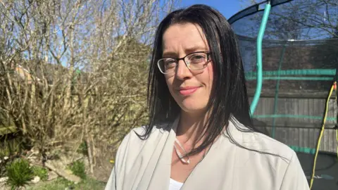 Chanel Walsh in the garden of her home. There is a trampoline behind her. She is smiling and looking at the camera.  She has long dark hair past her shoulders and wear half-rimmed glasses.