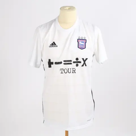 Bishop & Miller Auctioneers A general view of Ed Sheeran's Ipswich Town away shirt from the 2021/22 season. It is a white football shirt with the club's logo on the upper right hand side and the Adidas brand logo on the upper left hand side. Ed Sheeran's Divide album logo is in the middle of the shirt.