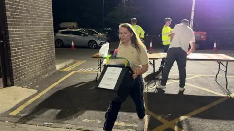 A woman is carrying a ballot box from a table in a parking lot into a building.