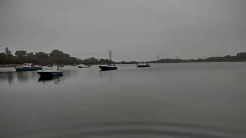 Stephles Eight small motorboats are moored in Hamworthy harbour. The water is grey and matches the colour of the sky. On the horizon you can see trees and electricty pylons on a gloomy day.