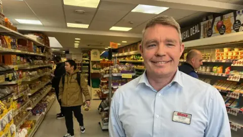 Zed Stott, a man with short, light-coloured hair, wearing a pale blue shirt with a name badge on his chest. He is standing in front of a supermarket aisle, with fridges and shelves, stacked with an assortment of sandwiches and bread.