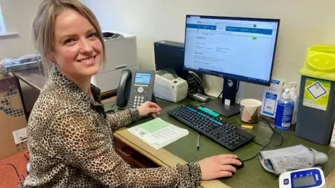 Dr Meleri Evans has light brown hair and is smiling at a camera. She is sat at a computer in a GP's office, wearing a leopard print shirt