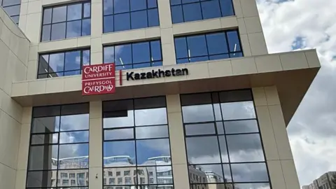 Cardiff University A large building with mirrored windows with a Cardiff University, Kazakhstan sign on the front and a row of Welsh, Kazakh, UK and Cardiff University flags along the front of the building.