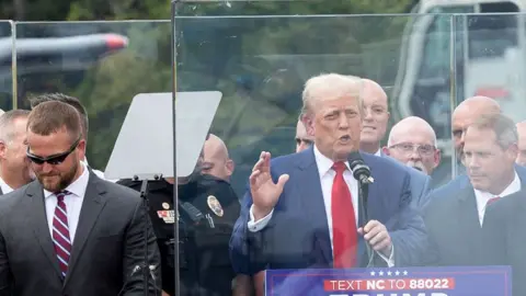 Donald Trump raises his hand while speaking alongside law enforcement while behind bulletproof glass at a rally in Asheboro on 21 August