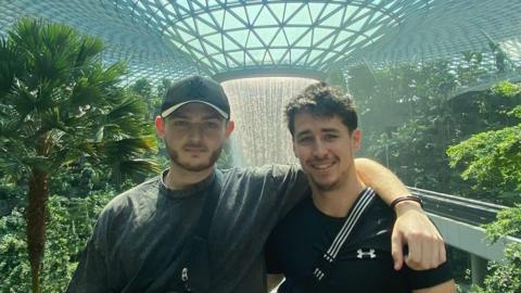 Toby Pearl and Liam James-Morris standing arm in arm in Singapore surrounded by an indoor garden and a dramatic water feature behind them.