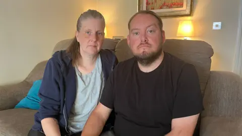 Gary and Sarah are sat on a sofa. Gary is wearing a black t-shirt and Sarah is wearing a grey t-shirt and navy jacket.