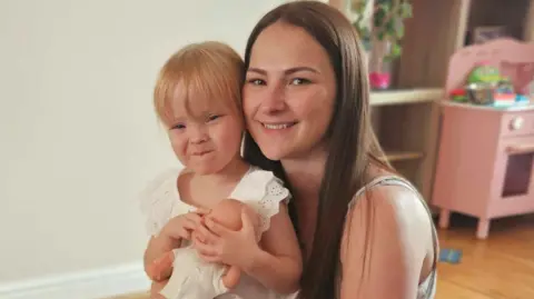 Family handout Hope smiles and is holding a baby doll while sitting on her mum Tanya's knee. Tanya is also smiling