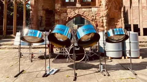 Impact Steel Band A row of steel drums pictured on a sunny day. A building with red bricks can be seen behind then.