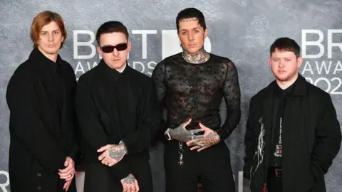Getty Images Bring Me The Horizon band on the Brit Awards red carpet. Left to right as we look are Matt Kean, Matt Nicholls, Oli Sykes and Lee Malia. All four men are wearing black. Matt Nicholls has black sunglasses on and tattoos visible on his hands. Oli also has tattoos covering both hands and is wearing an almost see-through black netting style top.