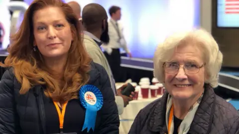 Wolverhampton Conservative Group Jane is wearing a navy jacket and has long red hair and is smiling at the camera. Her mother is on her right and is wearing glasses with short grey hair. She is also wearing a dark coloured jacket and scarf around her neck.
